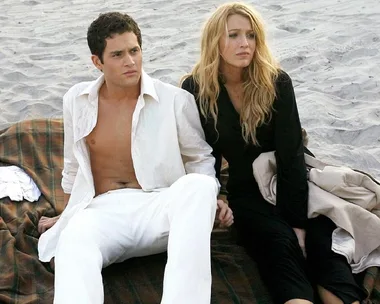 A couple sitting on a blanket at the beach, wearing casual white and black outfits, looking contemplative.