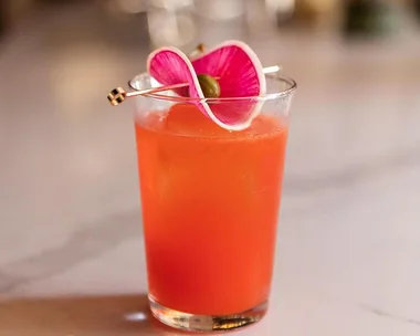 A vibrant orange cocktail garnished with a pink flower and olive on a metal skewer.