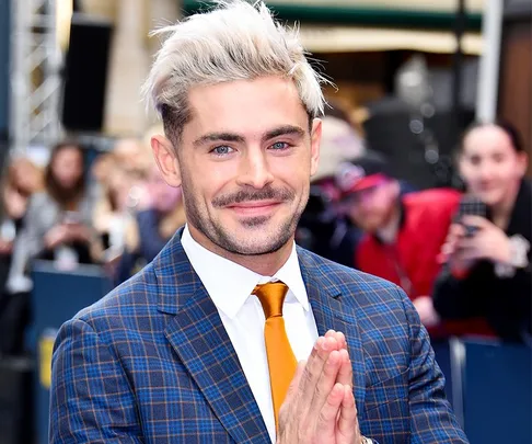 Person in a blue plaid suit with blond hair smiling at an event, with blurred crowd in the background.