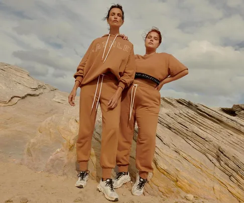 Two women stand on rocky terrain wearing matching brown athleisure outfits by AJE ATHLETICA under a cloudy sky.