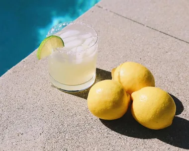 A margarita with a lime wedge on a poolside ledge next to three lemons.