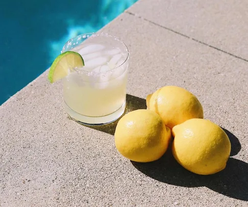 A margarita with a lime wedge on a poolside ledge next to three lemons.