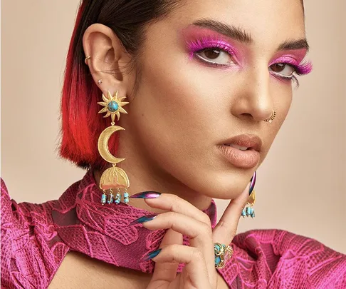 A woman with vibrant pink makeup and feathered lashes wearing crescent moon earrings and intricate jewelry.