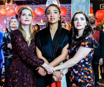 Three women holding hands, looking hopeful or inspired, surrounded by people and red lanterns in a festive setting.