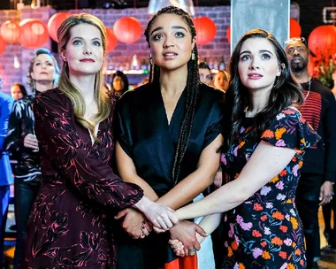 Three women holding hands, looking hopeful or inspired, surrounded by people and red lanterns in a festive setting.