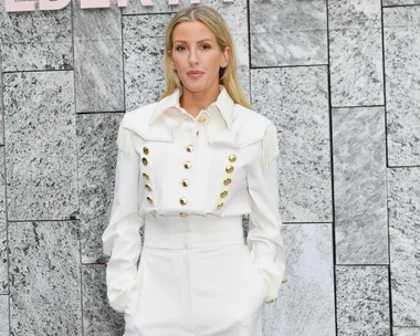 A person in a white outfit with gold buttons stands against a gray stone background.