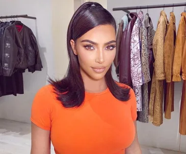 A woman in an orange top with sleek hair poses in a room with hanging jackets and coats.