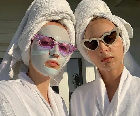 Two people wearing sheet masks, bathrobes, and sunglasses, with towels wrapped around their heads.