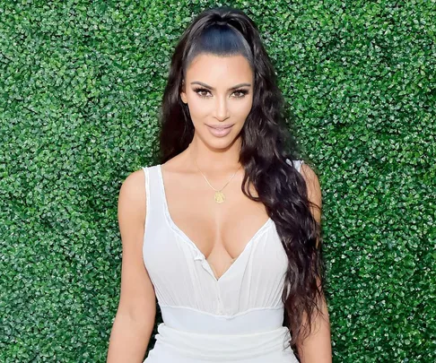 A woman with long dark hair in a white dress poses in front of a leafy green background.