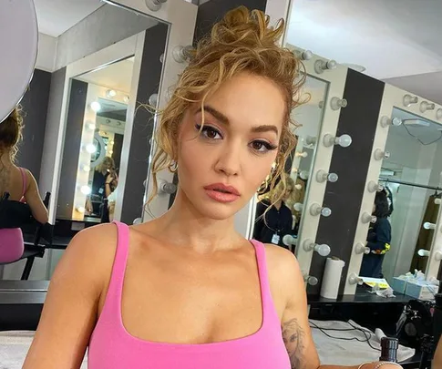 Person with curly hair in a pink top inside a makeup room with mirror lights.