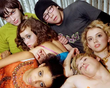 A group of young people from the TV show "Skins" lie together on a bed, looking at the camera.