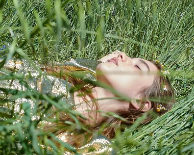 Woman in a sequined dress lying in a lush green meadow, eyes closed, surrounded by tall grass and wildflowers.