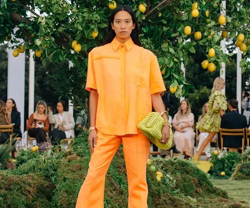 Woman in bright orange outfit holding green clutch, standing in garden setting with lemon tree and people seated in background.