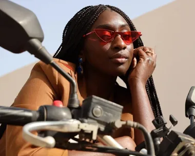 A woman in red sunglasses, resting on a motorcycle handlebar, wearing a brown jacket with braided hair.