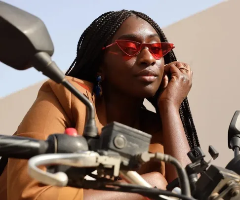 A woman in red sunglasses, resting on a motorcycle handlebar, wearing a brown jacket with braided hair.