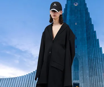 Person in oversized black jacket and cap in front of a large, modern tower building.