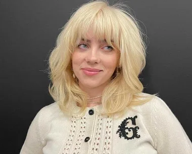 A person with blonde hair wearing a white top and black background.