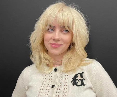 A person with blonde hair wearing a white top and black background.