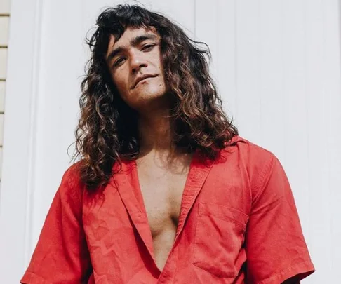 Man with long hair wearing a red shirt, standing against a white background.