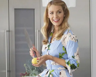 Woman in floral dress smiling, holding a knife and a lemon in a modern kitchen setting.