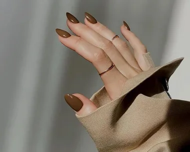 A hand with chocolate-colored nails and gold rings, wearing a beige sleeve, against a light background.