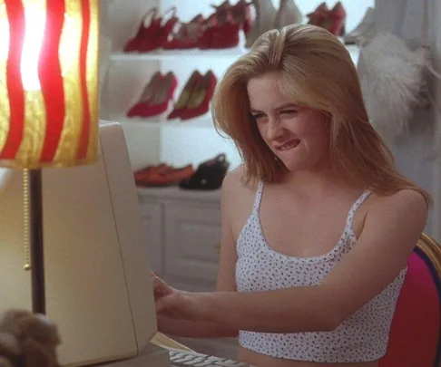 Young woman in a white crop top using a computer, surrounded by colorful shoes on shelves.