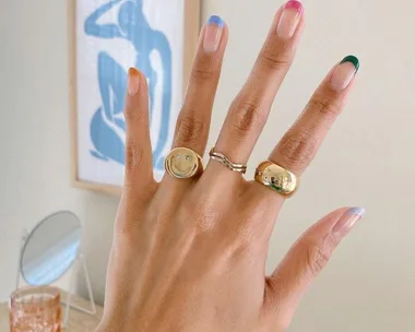 A hand with a colorful French manicure, wearing three gold rings, against a background with blue abstract art.