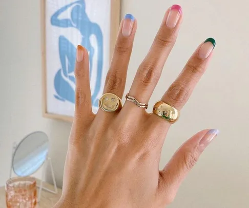 A hand with a colorful French manicure, wearing three gold rings, against a background with blue abstract art.