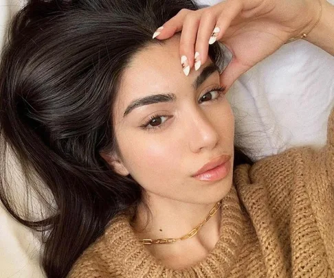 A person with long dark hair and a knitted sweater, lying down with hand on forehead, showing neat nails.