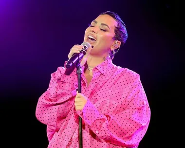 Person singing passionately on stage, wearing a pink polka dot shirt, lit by purple stage lighting.