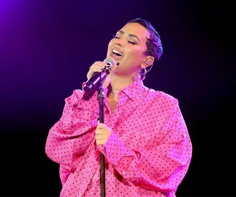 Person singing passionately on stage, wearing a pink polka dot shirt, lit by purple stage lighting.