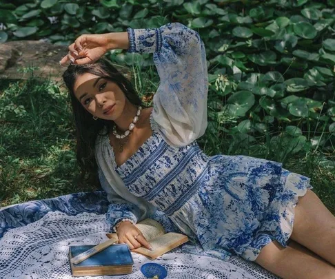 Woman in a blue and white floral dress lies on a picnic blanket, surrounded by books and greenery.