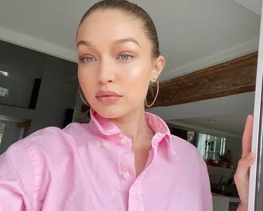 A woman with light brown hair tied up, wearing a pink shirt and hoop earrings, stands indoors, looking into the camera.