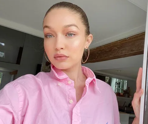 A woman with light brown hair tied up, wearing a pink shirt and hoop earrings, stands indoors, looking into the camera.