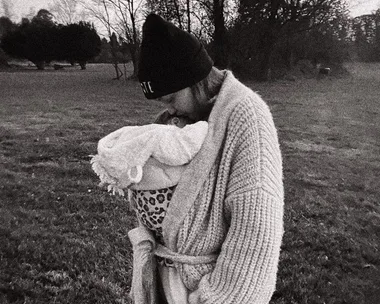 Black and white photo of person in knit sweater and beanie holding a baby in a field.