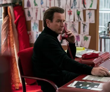 Man in black turtleneck on red phone at a desk with fashion sketches on the wall behind him.