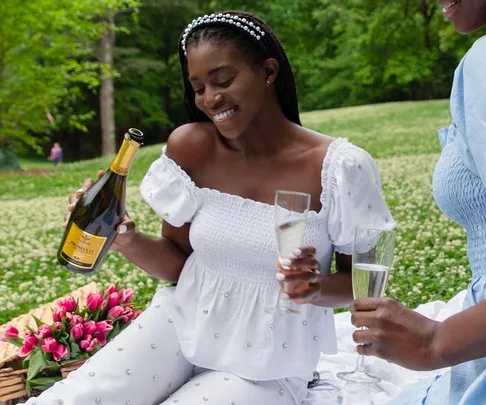 Woman in white dress smiles, holding champagne and glass at picnic with flowers in a green park.