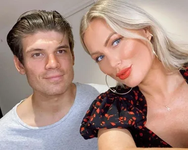 A couple smiling for a selfie indoors, with a woman in a patterned top and a man in a grey shirt.