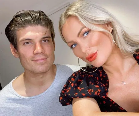 A couple smiling for a selfie indoors, with a woman in a patterned top and a man in a grey shirt.