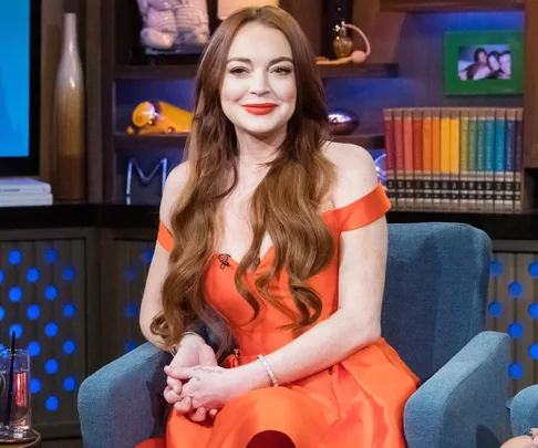 A woman with long red hair in an orange dress sits on a blue chair, smiling, with books on shelves behind her.