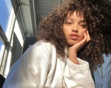 A person with curly hair in a white robe lit by sunlight indoors.