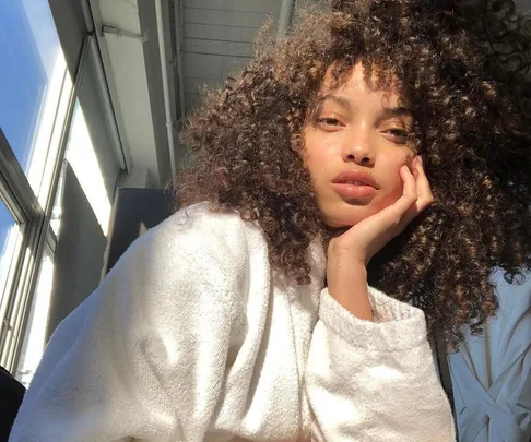 A person with curly hair in a white robe lit by sunlight indoors.