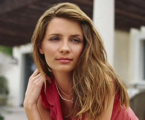 Young woman with long hair in a red top, outdoors with soft lighting and neutral expression.