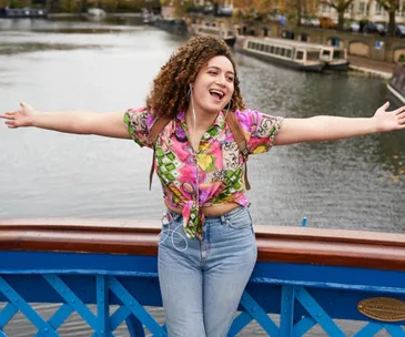 A woman joyfully posing on a blue bridge near water, wearing a colorful shirt and jeans, with arms outstretched.