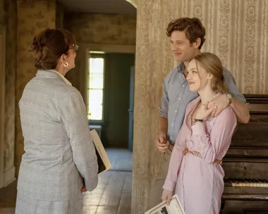 A couple stands together in a room with a piano, speaking with a woman holding a document.