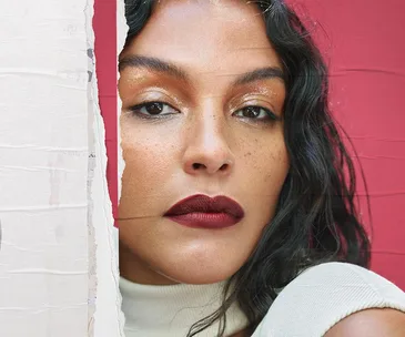 Close-up of a model with glossy eye makeup and dark red lipstick against a pink background.