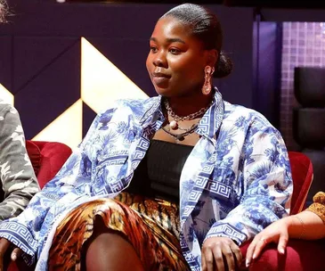 A person sitting on a red chair wearing a blue patterned shirt, black top, and animal print skirt.