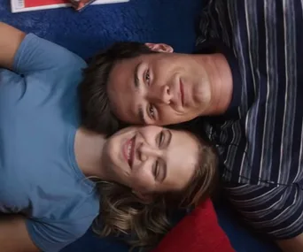 A couple smiling while lying on their backs, heads touching, one in a blue shirt and the other in a striped shirt.