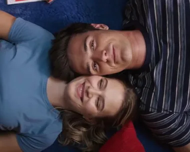 A couple smiling while lying on their backs, heads touching, one in a blue shirt and the other in a striped shirt.