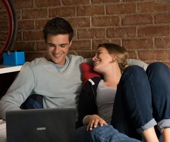 A couple on a couch, smiling at a laptop, in front of a brick wall.